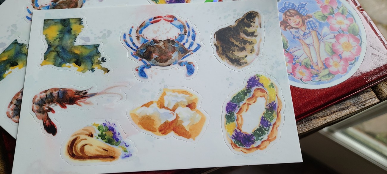 A sticker sheet with 7 different designs- 2 kingcake, a blue crab, an oyster, a shrimp, and beignets.